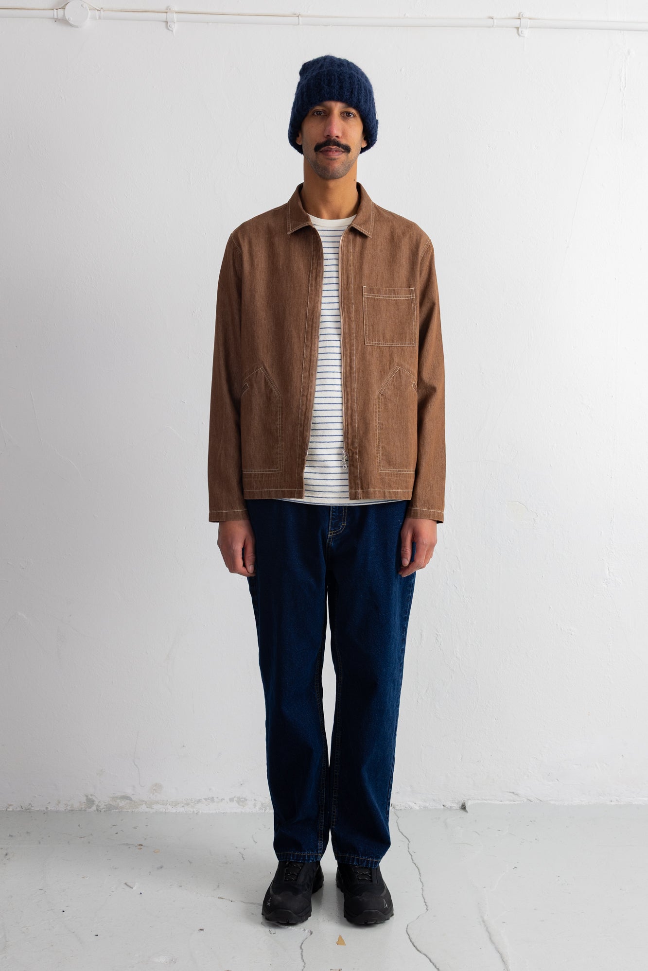 A man wearing a brown suede jacket over a white striped t-shirt, dark blue jeans, black shoes, and a navy blue beanie, standing against a white wall.
