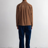 A person standing with their back to the camera, wearing a brown corduroy jacket, dark blue jeans, a navy blue beanie, and black shoes, set against a plain white wall.