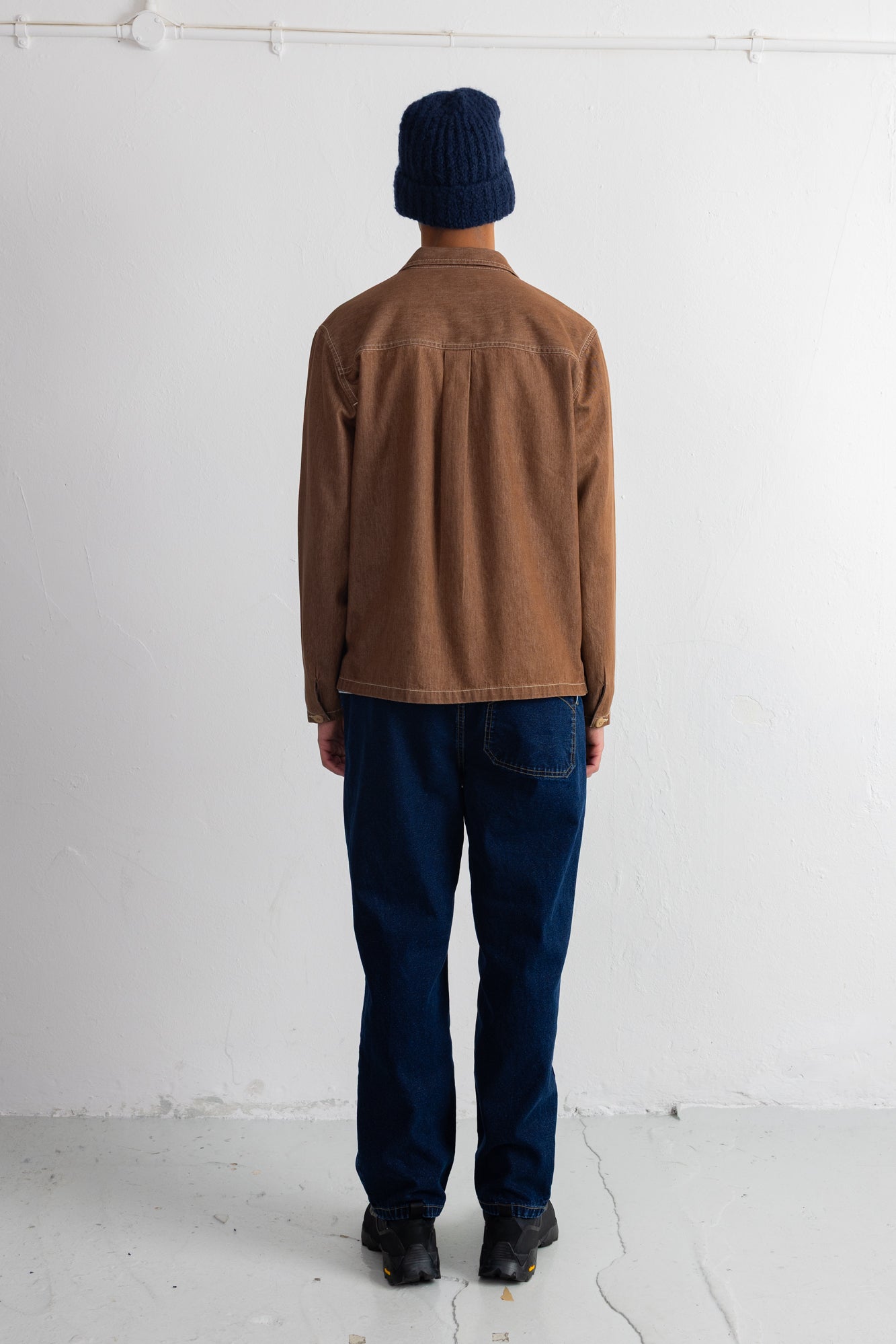 A person standing with their back to the camera, wearing a brown corduroy jacket, dark blue jeans, a navy blue beanie, and black shoes, set against a plain white wall.