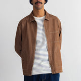 A man wearing a brown corduroy utility jacket over a white and navy striped t-shirt, paired with a navy blue beanie and dark blue jeans, standing against a plain white wall.