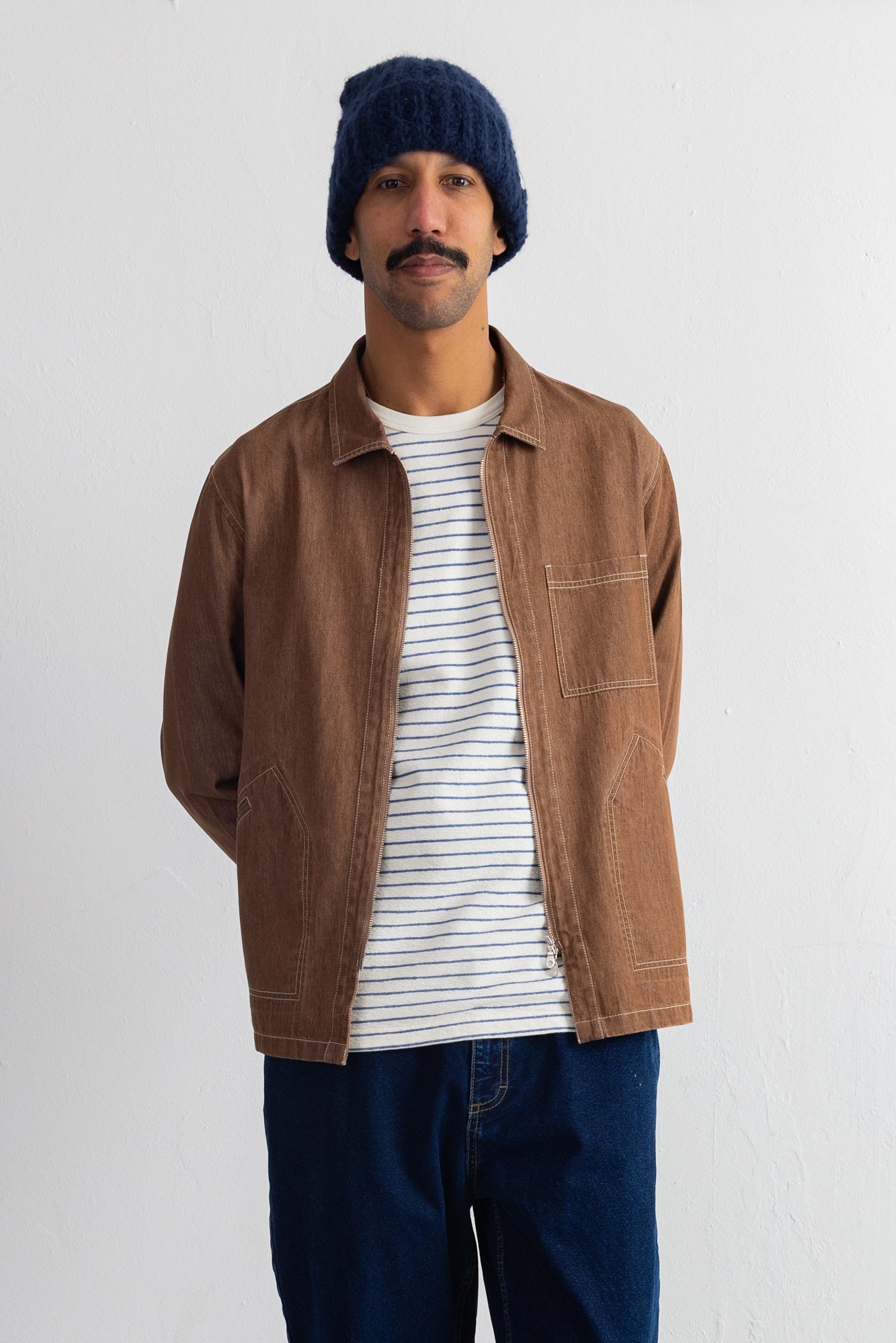 A man wearing a brown corduroy utility jacket over a white and navy striped t-shirt, paired with a navy blue beanie and dark blue jeans, standing against a plain white wall.