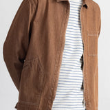 A man wearing a brown canvas utility jacket over a white t-shirt with thin blue stripes and blue jeans, photographed against a plain white background.