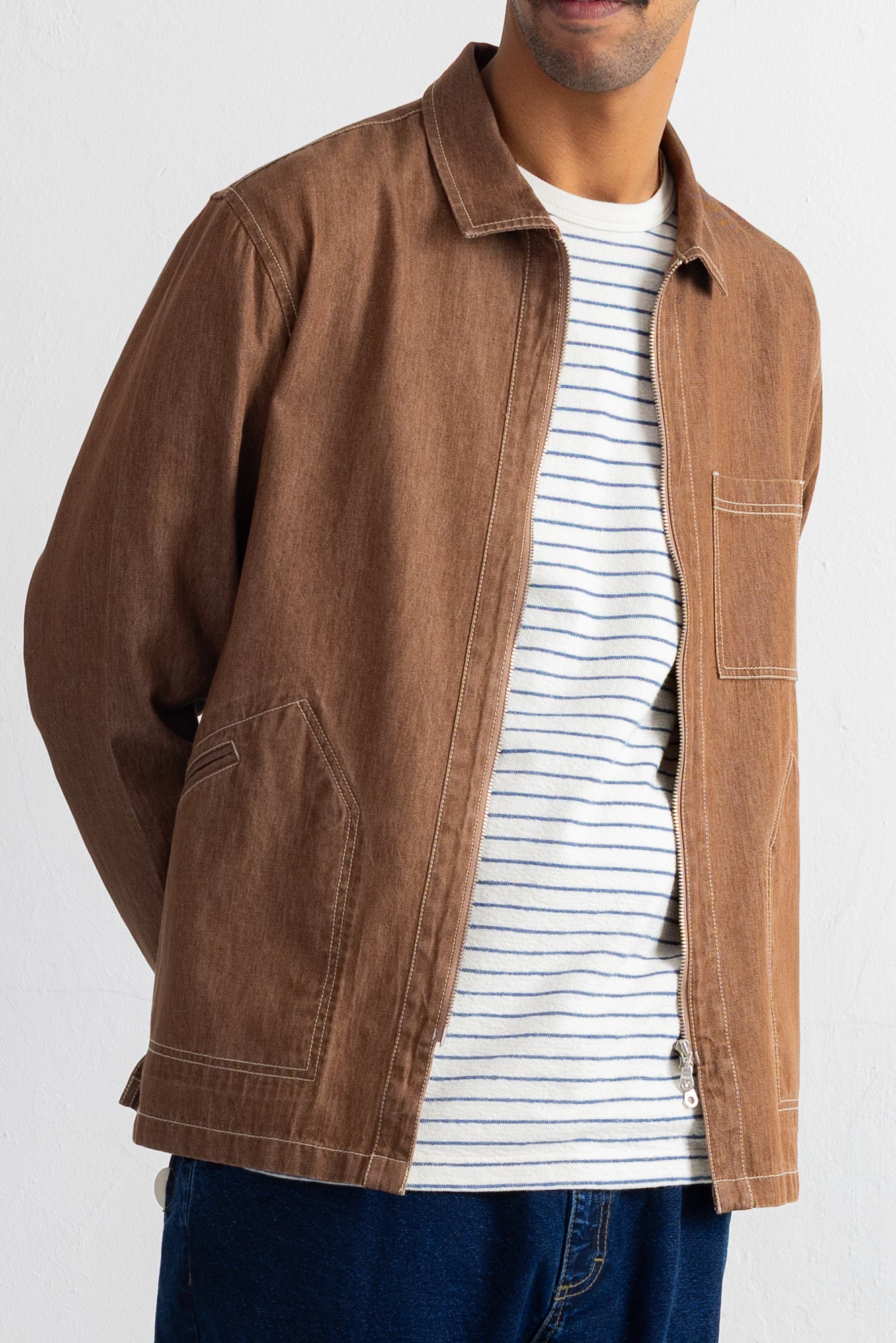 A man wearing a brown canvas utility jacket over a white t-shirt with thin blue stripes and blue jeans, photographed against a plain white background.