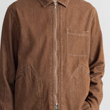 A man wearing a brown zip-up utility jacket with contrast stitching, a chest pocket, and two large lower patch pockets, paired with blue jeans against a plain white wall.