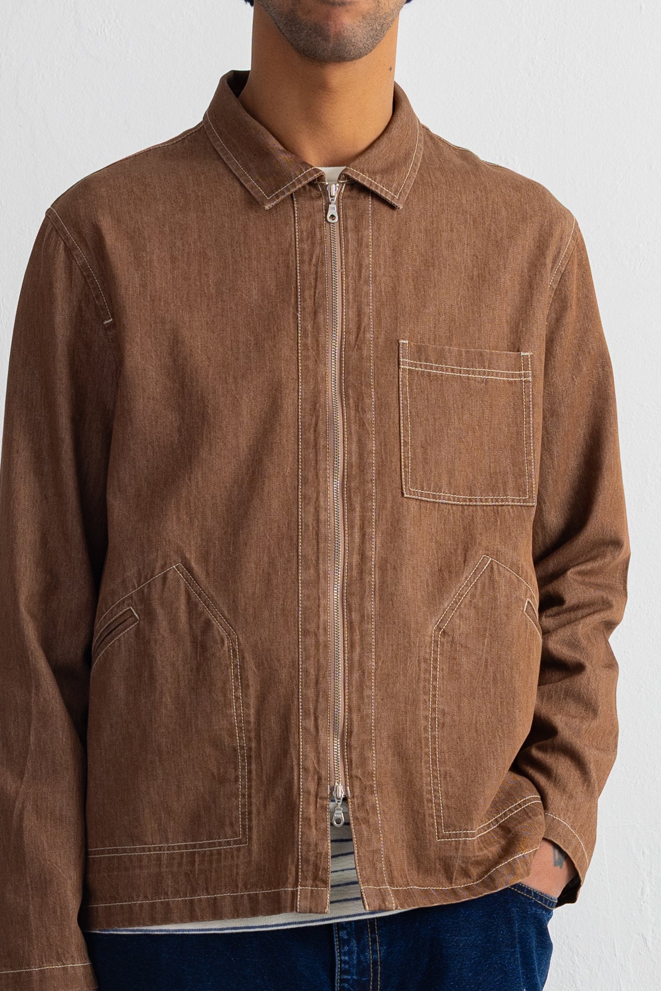 A man wearing a brown zip-up utility jacket with contrast stitching, a chest pocket, and two large lower patch pockets, paired with blue jeans against a plain white wall.