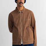 A man wearing a brown zip-up jacket, a navy blue knit beanie, and dark blue jeans, standing against a plain off-white wall.