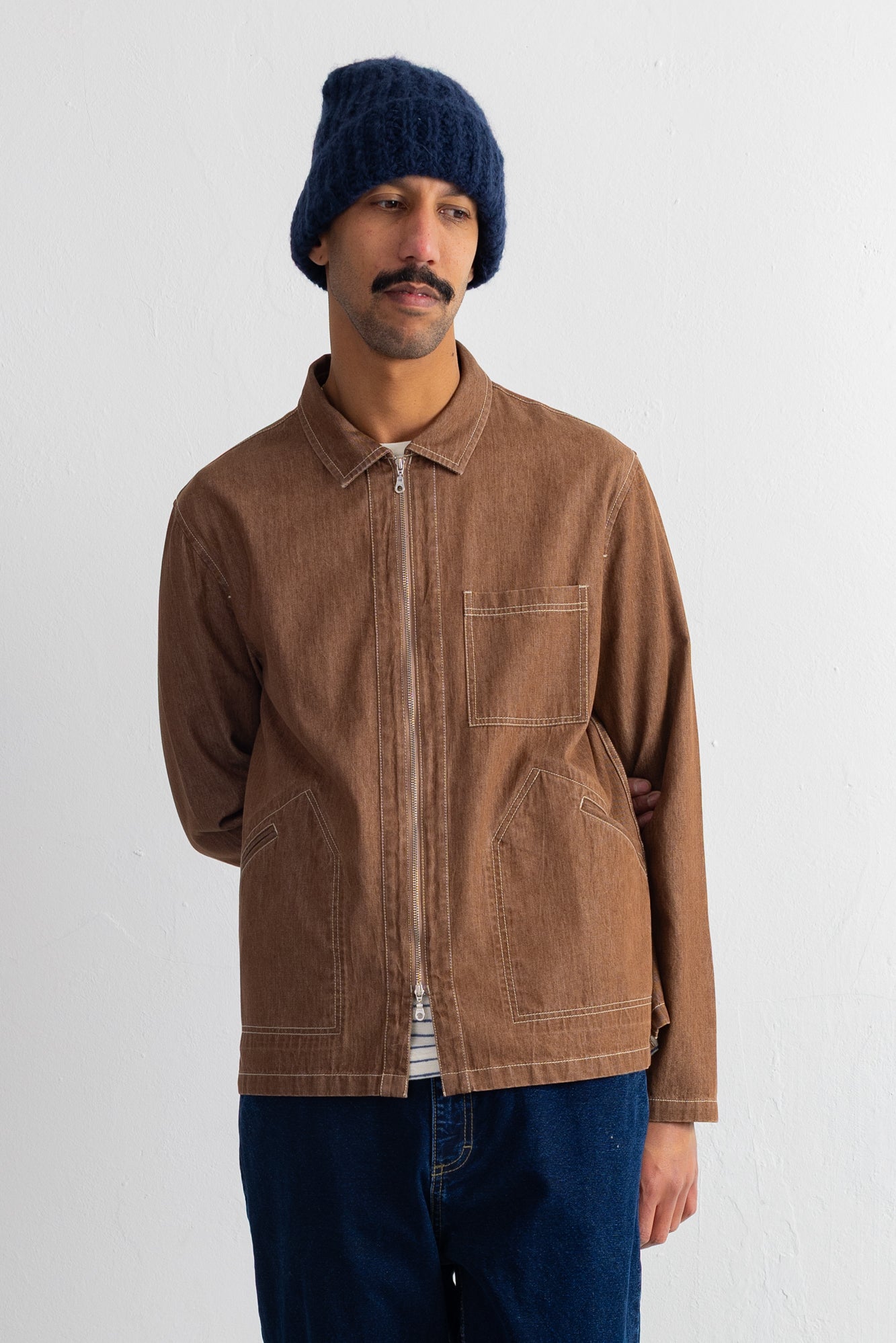 A man wearing a brown zip-up jacket, a navy blue knit beanie, and dark blue jeans, standing against a plain off-white wall.