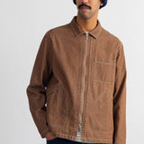 A man wearing a brown zip-up canvas jacket, a navy blue knit beanie, and dark blue jeans, standing against a plain white studio background.