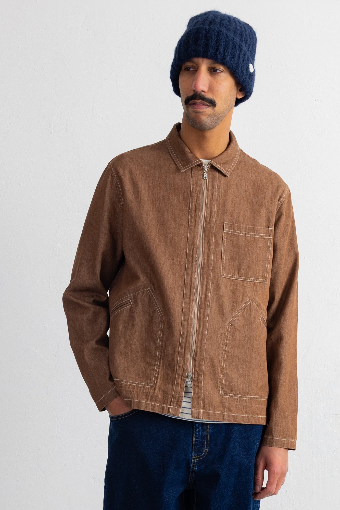 A man wearing a brown zip-up canvas jacket, a navy blue knit beanie, and dark blue jeans, standing against a plain white studio background.