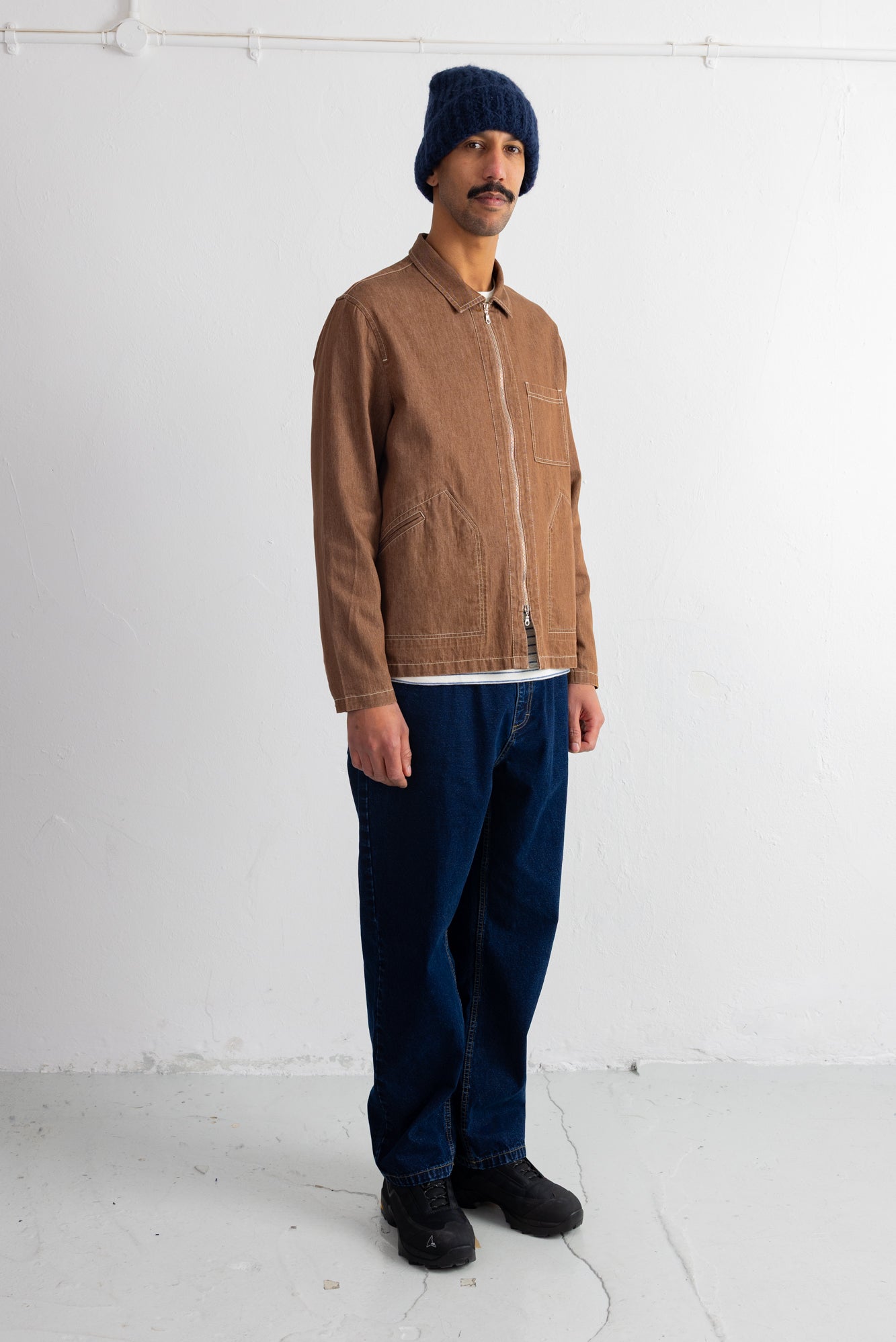 A man wearing a brown corduroy jacket, dark blue jeans, a navy blue beanie, and black shoes, standing against a white studio wall.