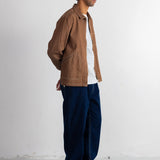 A man wearing a brown canvas utility jacket over a white and navy striped shirt, dark blue jeans, a navy beanie, and black shoes, standing in profile against a white wall.