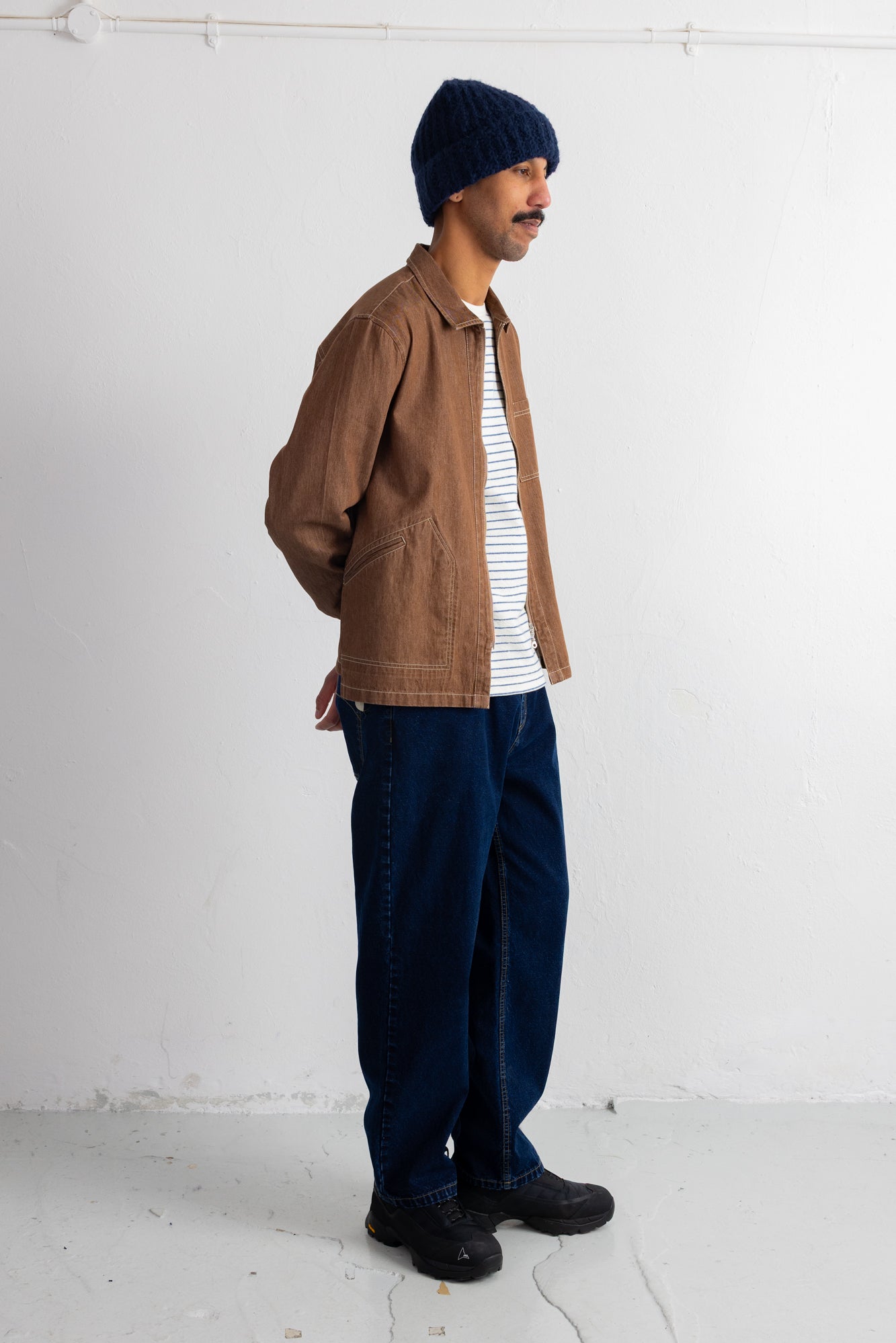 A man wearing a brown canvas utility jacket over a white and navy striped shirt, dark blue jeans, a navy beanie, and black shoes, standing in profile against a white wall.