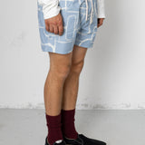 Person wearing blue shorts with white geometric pattern, white shirt, maroon socks, and black shoes on a light gray background.