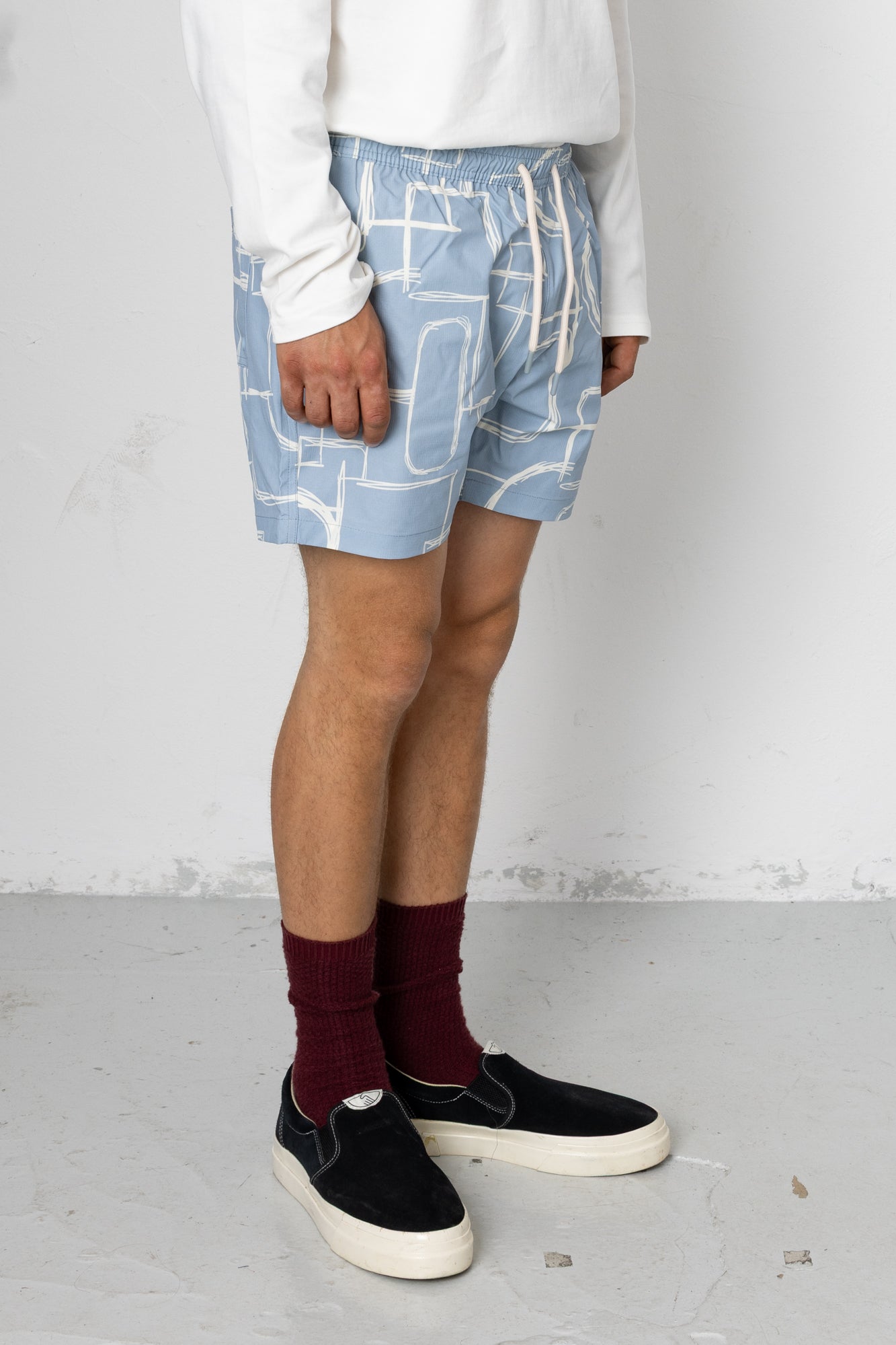 Person wearing blue shorts with white geometric pattern, white shirt, maroon socks, and black shoes on a light gray background.