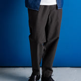 A person wearing dark charcoal gray relaxed-fit trousers, styled with a white t-shirt, a blue denim shirt, and black leather shoes, standing against a vibrant blue studio backdrop.