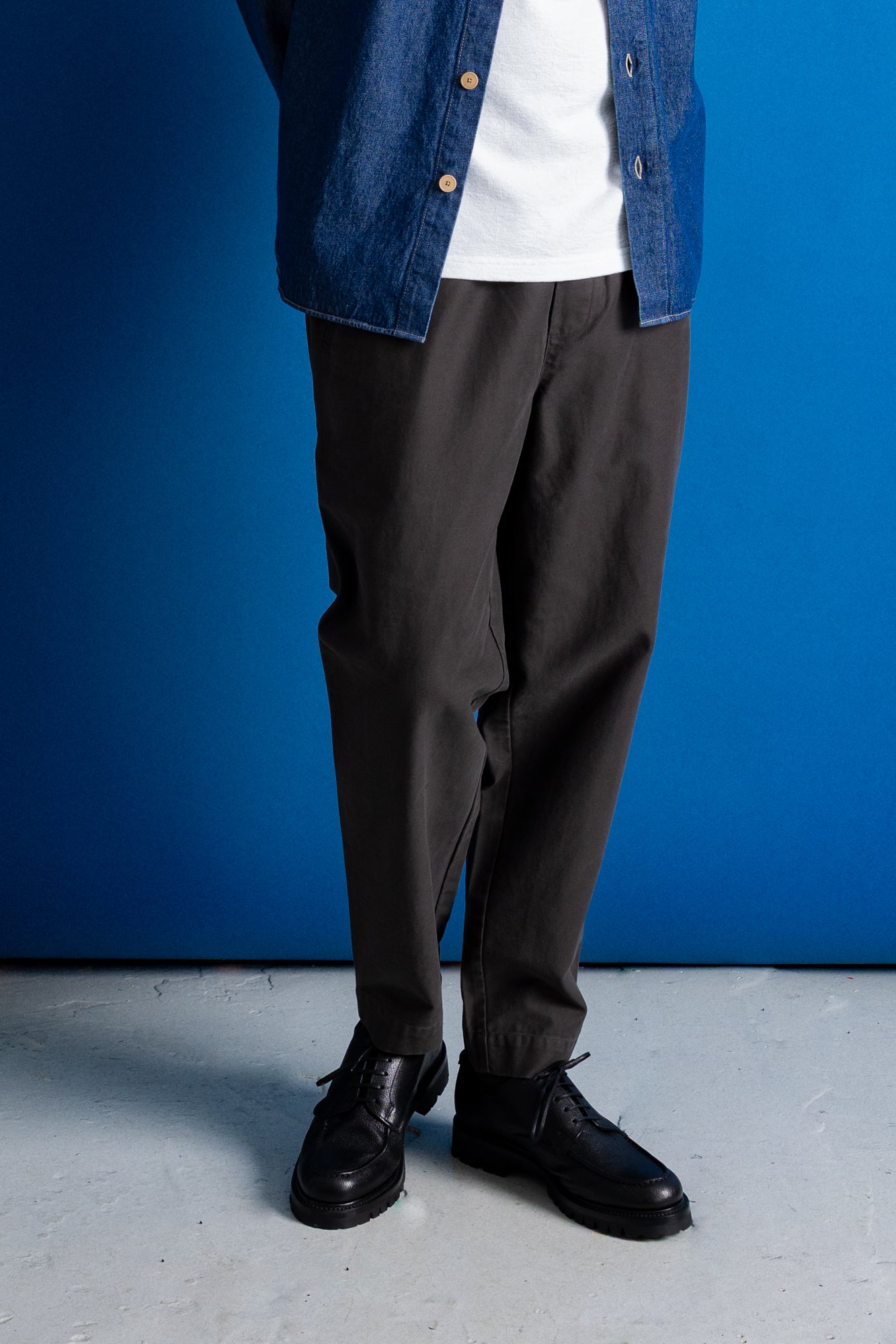 A person wearing dark charcoal gray relaxed-fit trousers, styled with a white t-shirt, a blue denim shirt, and black leather shoes, standing against a vibrant blue studio backdrop.