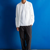A man stands against a bright blue studio backdrop wearing a white long-sleeve button-up shirt over a white undershirt, dark charcoal trousers, and black dress shoes.