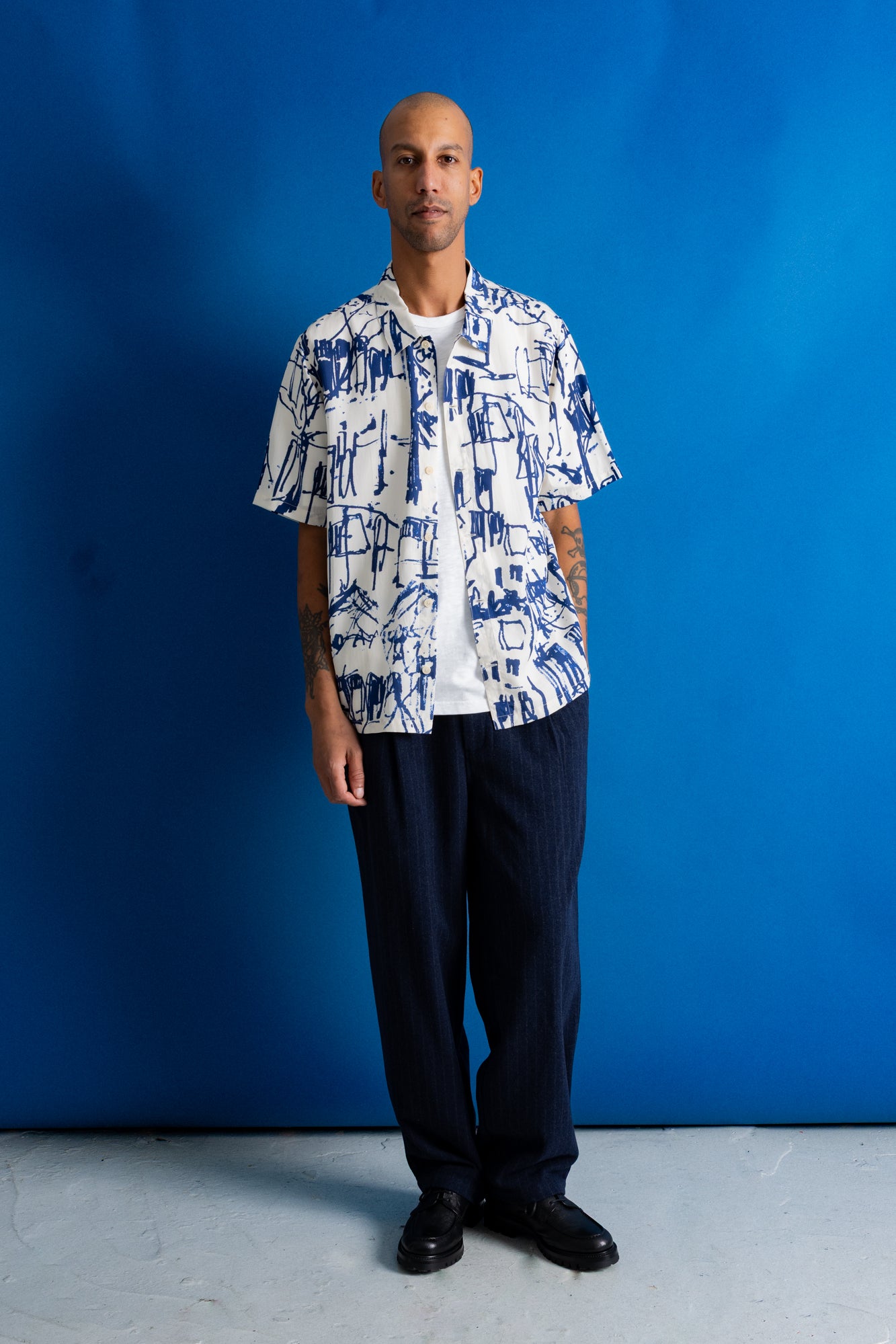 A man stands against a bright blue studio backdrop wearing a white short-sleeve button-up shirt with a blue abstract graphic print, dark navy trousers, and black shoes.