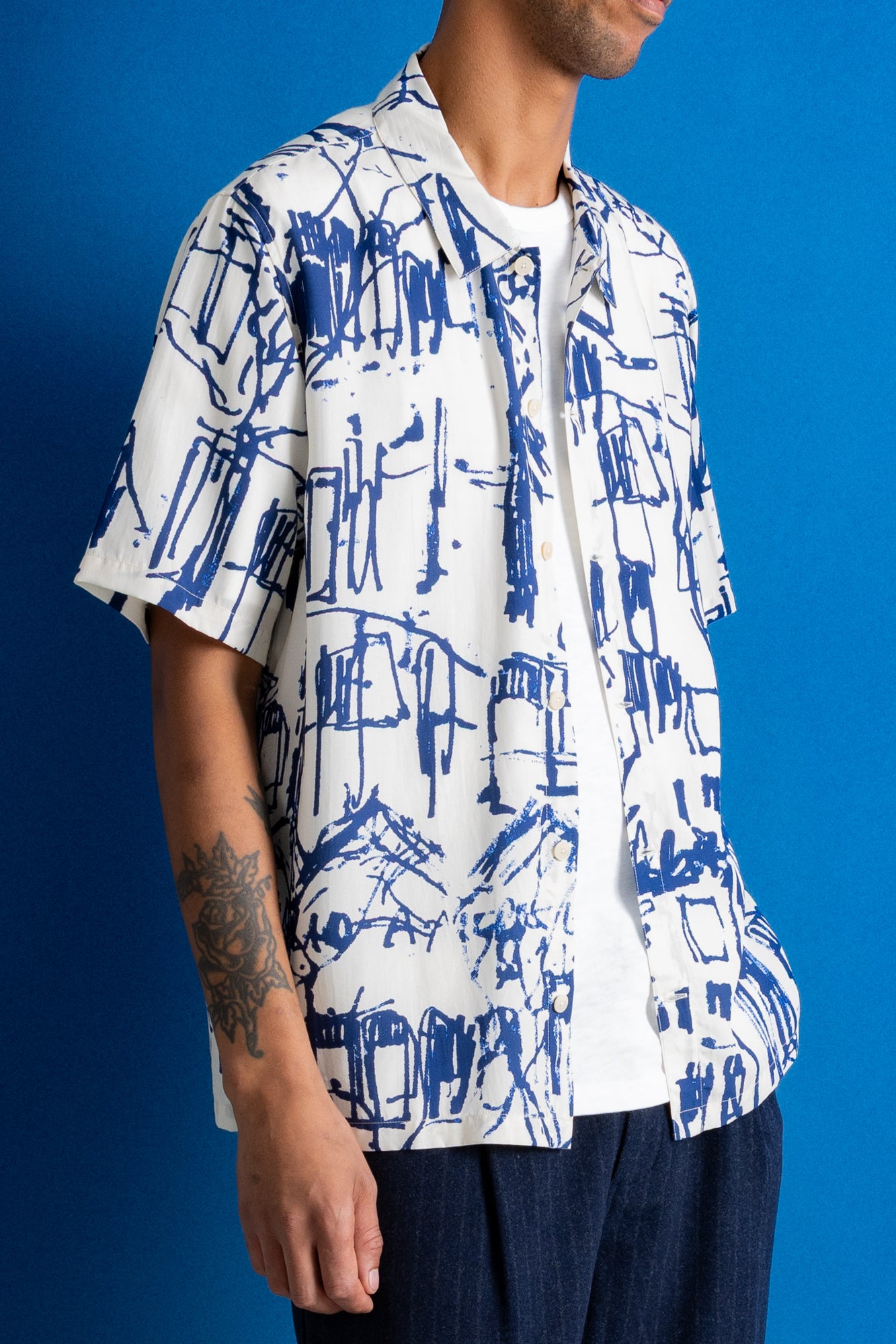 A man stands against a bright blue studio backdrop wearing a white short-sleeve button-up shirt with a blue abstract graphic print and dark navy trousers.