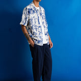 A man stands in profile wearing a white short-sleeve button-up shirt with a blue abstract line print, dark navy pinstriped trousers, and black shoes, posed against a solid blue studio backdrop.