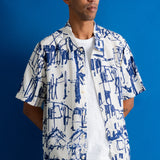 A man wearing a white short-sleeve button-up shirt with a blue abstract architectural print, layered over a white t-shirt and dark pants, standing against a solid blue background.