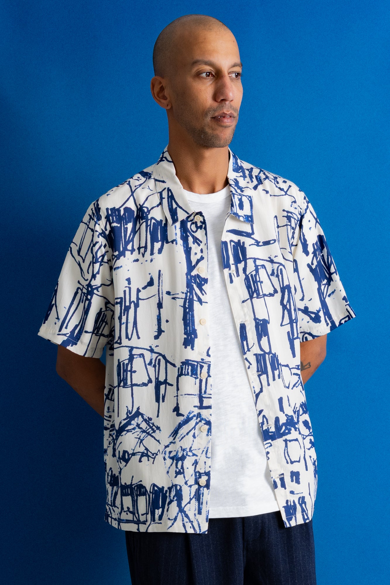 A man wearing a white short-sleeve button-up shirt with a blue abstract architectural print, layered over a white t-shirt and dark pants, standing against a solid blue background.