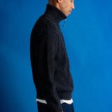 Man standing profile wearing zipped up high neck grey quarter zip jumper, white t-shirt and navy trousers against a blue backdrop.