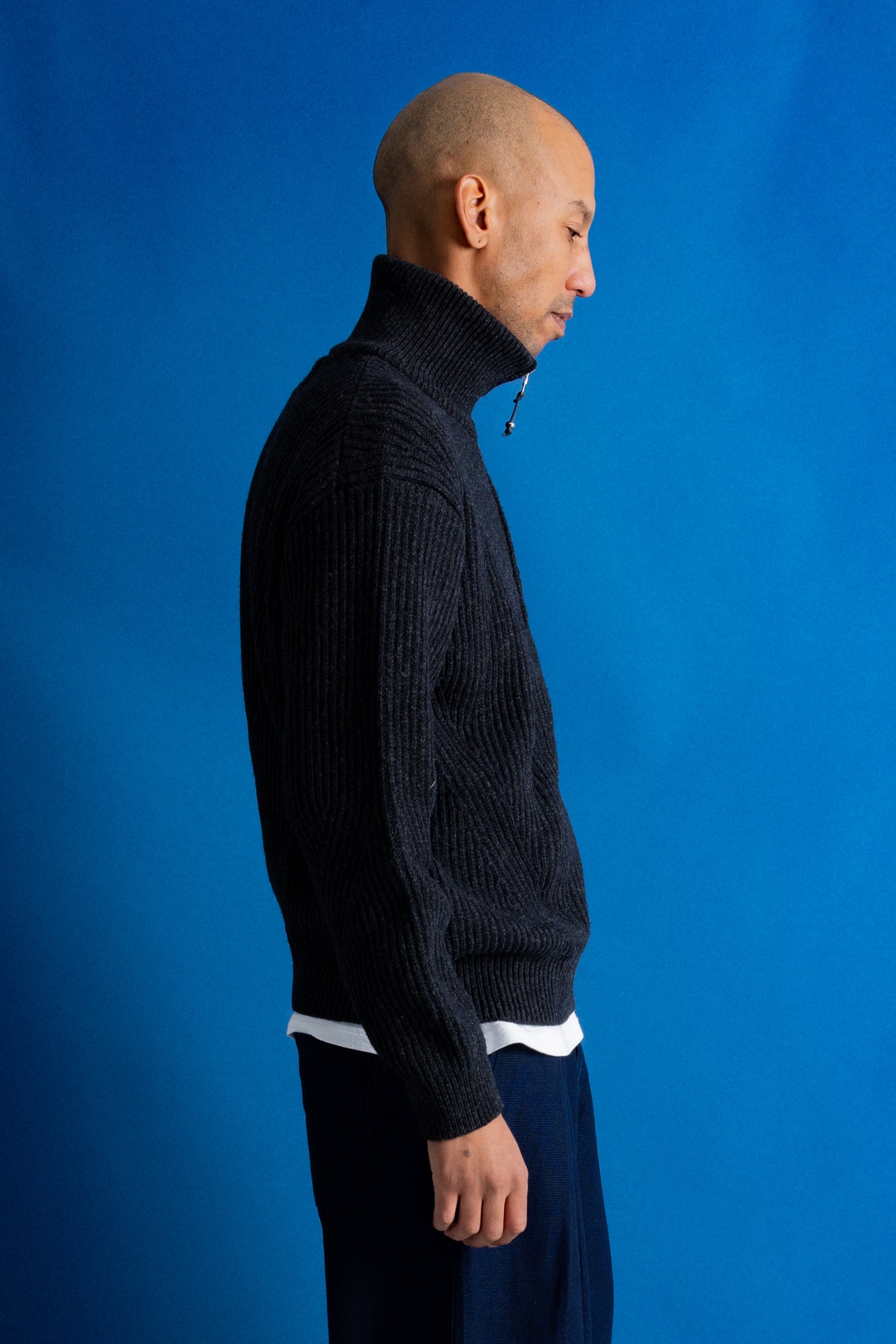 Man standing profile wearing zipped up high neck grey quarter zip jumper, white t-shirt and navy trousers against a blue backdrop.