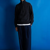 Man wearing grey quarter zip jumper, white t-shirt and navy trousers against a blue backdrop.