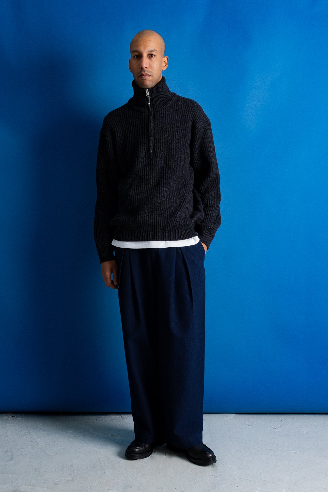 Man wearing grey quarter zip jumper, white t-shirt and navy trousers against a blue backdrop.