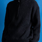 Man wearing zipped up high neck grey quarter zip jumper, white t-shirt and navy trousers against a blue backdrop.