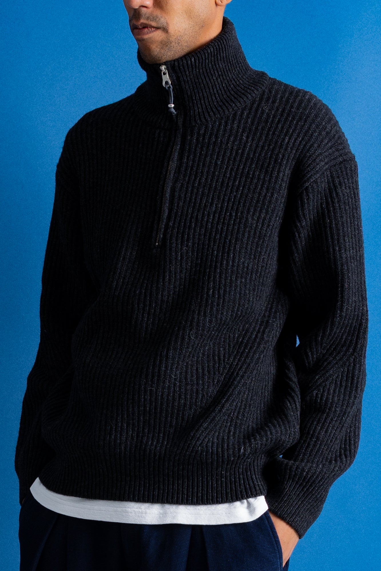 Man wearing zipped up high neck grey quarter zip jumper, white t-shirt and navy trousers against a blue backdrop.