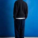 Man facing away from the camera wearing zipped up high neck grey quarter zip jumper, white t-shirt and navy trousers against a blue backdrop.