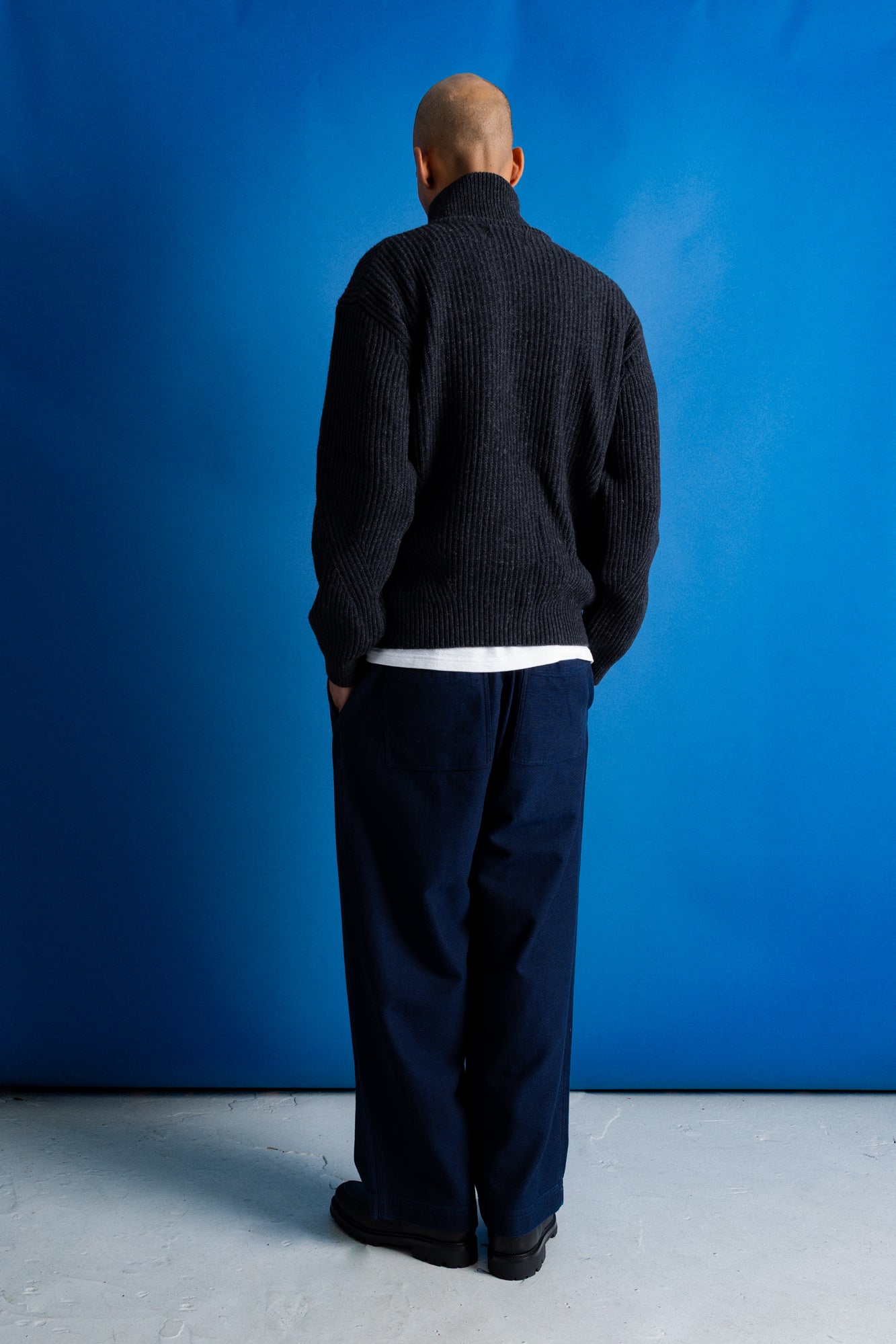 Man facing away from the camera wearing zipped up high neck grey quarter zip jumper, white t-shirt and navy trousers against a blue backdrop.