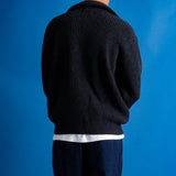 Man facing away from the camera wearing zipped up high neck grey quarter zip jumper, white t-shirt and navy trousers against a blue backdrop.