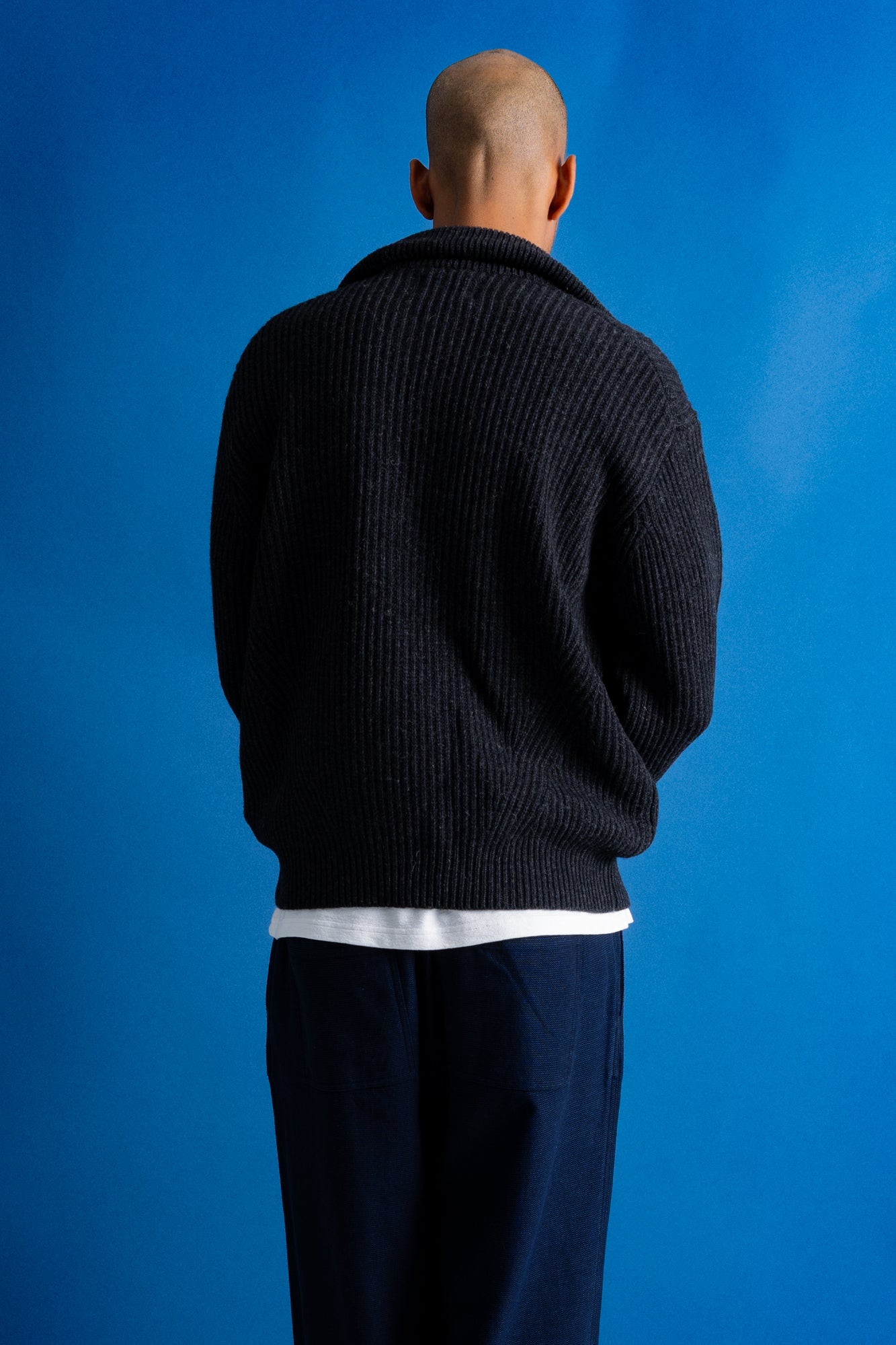 Man facing away from the camera wearing zipped up high neck grey quarter zip jumper, white t-shirt and navy trousers against a blue backdrop.