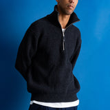 Man wearing grey quarter zip jumper, white t-shirt and navy trousers against a blue backdrop.