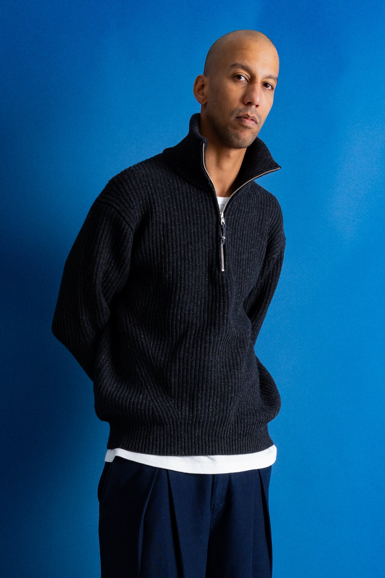 Man wearing grey quarter zip jumper, white t-shirt and navy trousers against a blue backdrop.