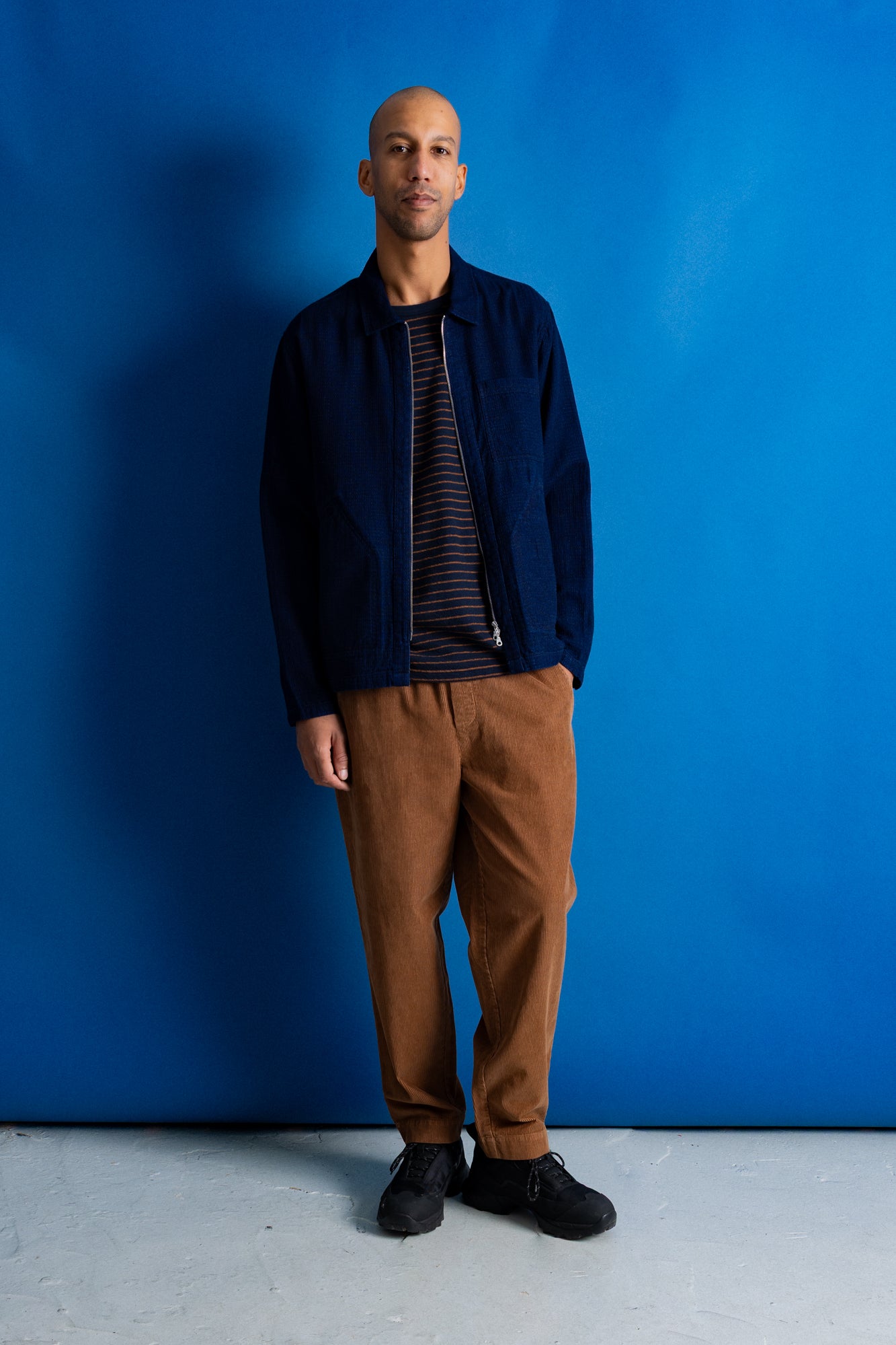 A man stands against a bright blue studio backdrop wearing a navy overshirt over a dark striped shirt, brown corduroy pants, and black shoes.