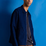A man wearing a dark blue textured zip-up jacket over a navy and orange striped t-shirt and brown corduroy pants, standing against a solid blue background.