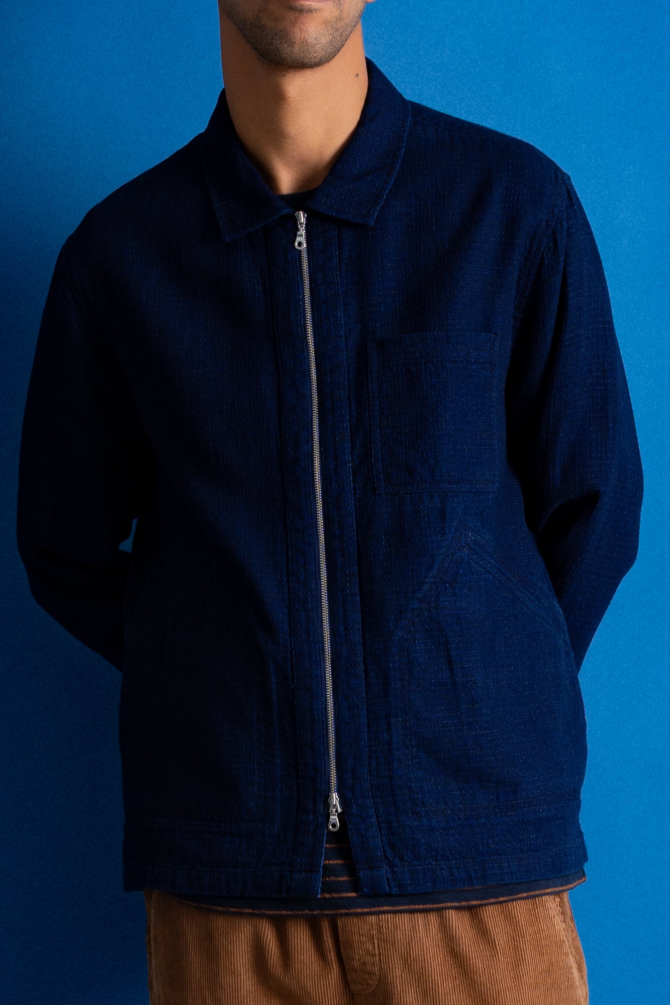 Man wearing a dark blue jacket, striped t-shirt and dark orange cord trousers against a bright blue background.
