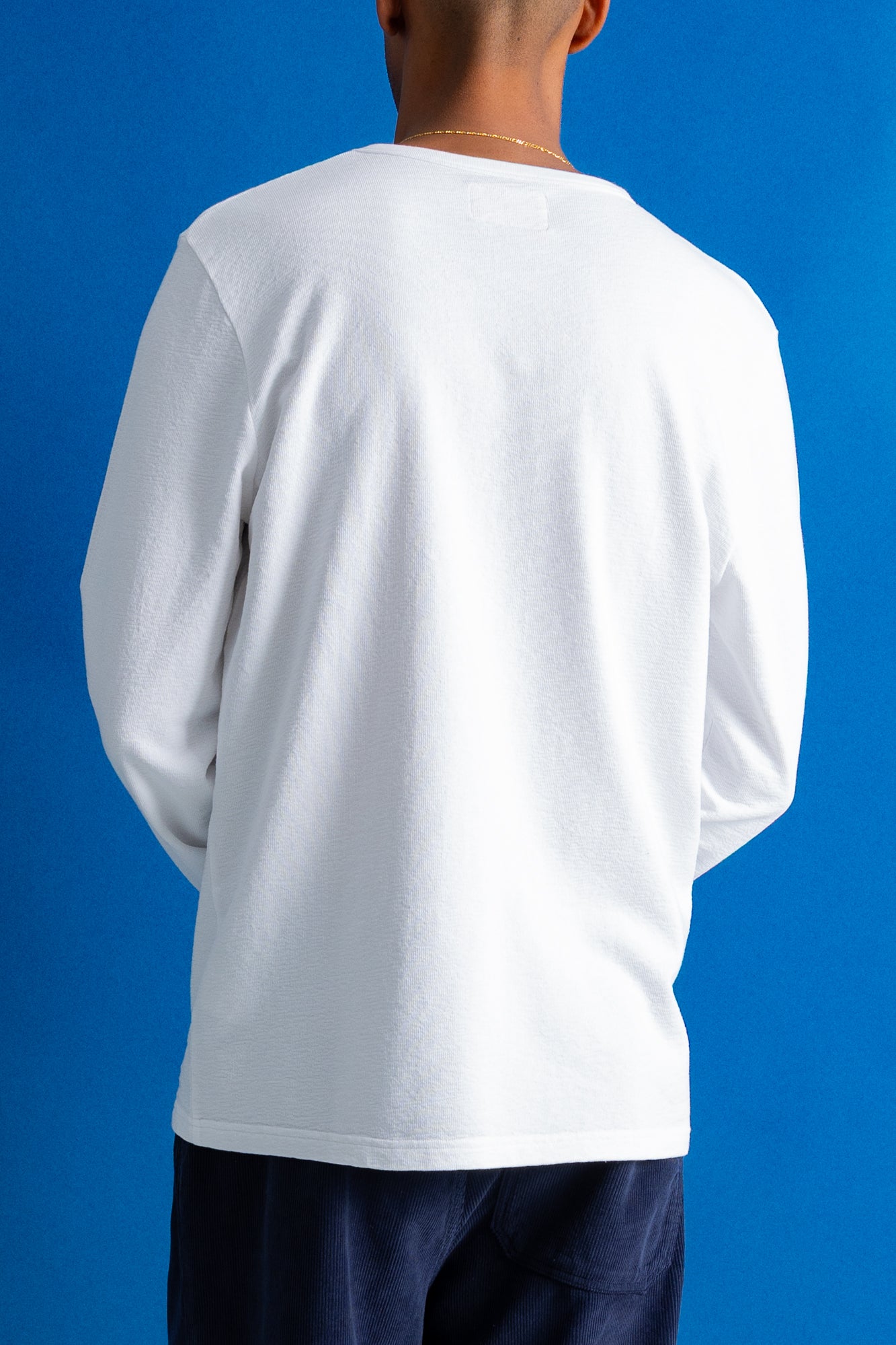 A person shown from the back wearing a plain white long-sleeve t-shirt and dark navy pants, standing against a solid blue background.