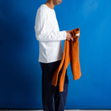 A man in a white long-sleeve shirt, dark trousers, and black shoes stands against a bright blue studio backdrop, holding up an orange ribbed knit sweater.