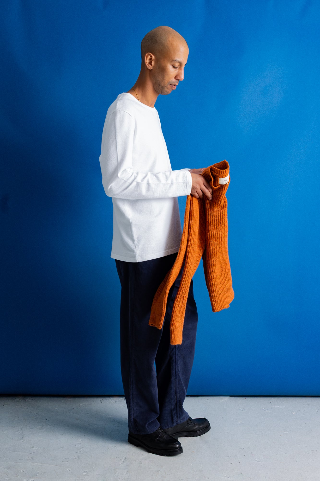 A man in a white long-sleeve shirt, dark trousers, and black shoes stands against a bright blue studio backdrop, holding up an orange ribbed knit sweater.