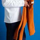 A person wearing a white long-sleeve shirt and dark navy pants holds an orange ribbed knit sweater against a solid blue background.