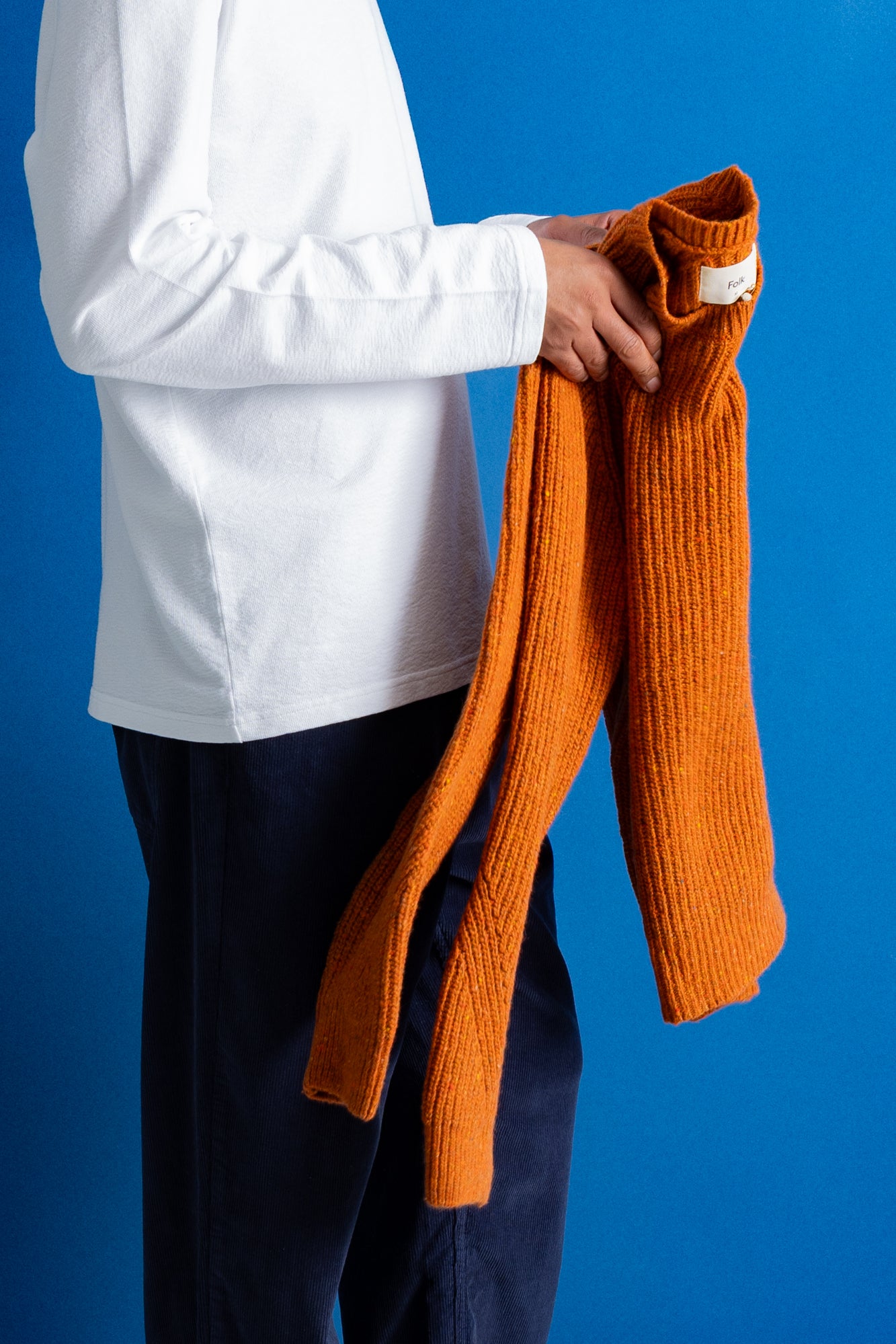A person wearing a white long-sleeve shirt and dark navy pants holds an orange ribbed knit sweater against a solid blue background.