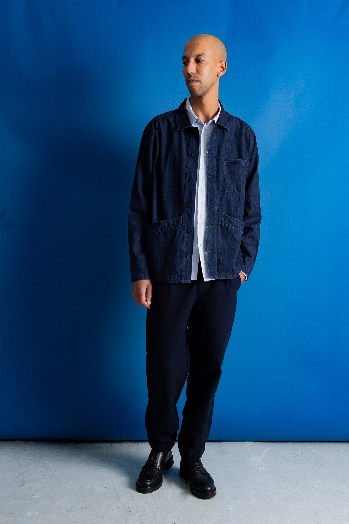 A man wearing a dark indigo denim utility jacket over a white collared shirt, paired with dark trousers and black leather shoes, standing against a vibrant blue studio backdrop.