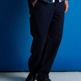 A person wearing dark navy trousers, a dark denim jacket over a white shirt, and black leather shoes, standing against a solid blue wall.