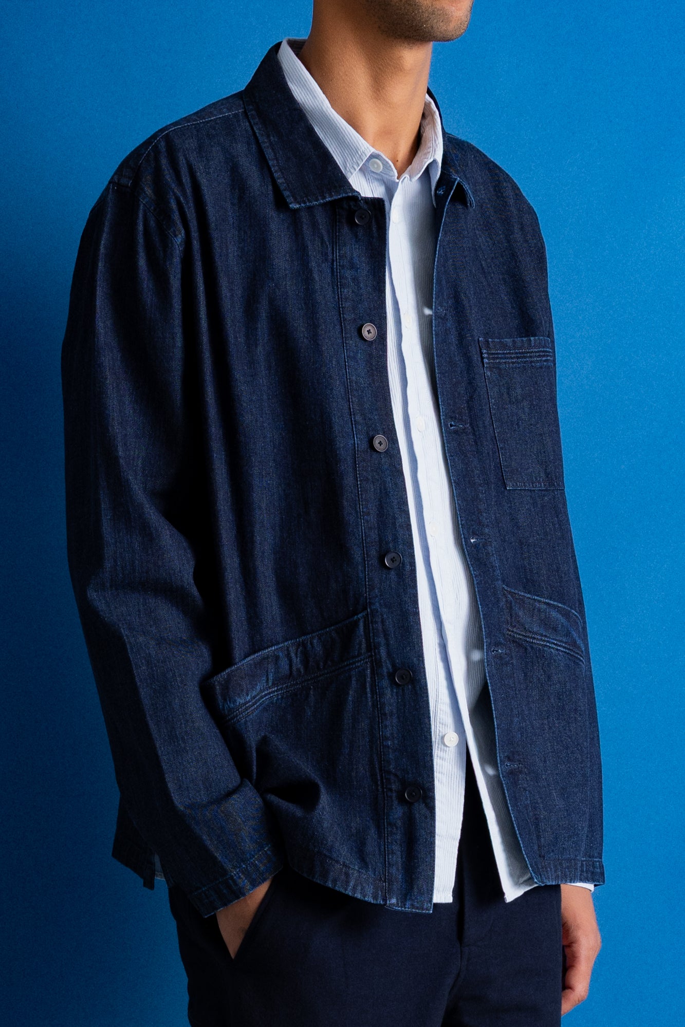 A man wearing a dark indigo denim utility jacket over a white collared shirt, shown from the neck down against a solid blue background.