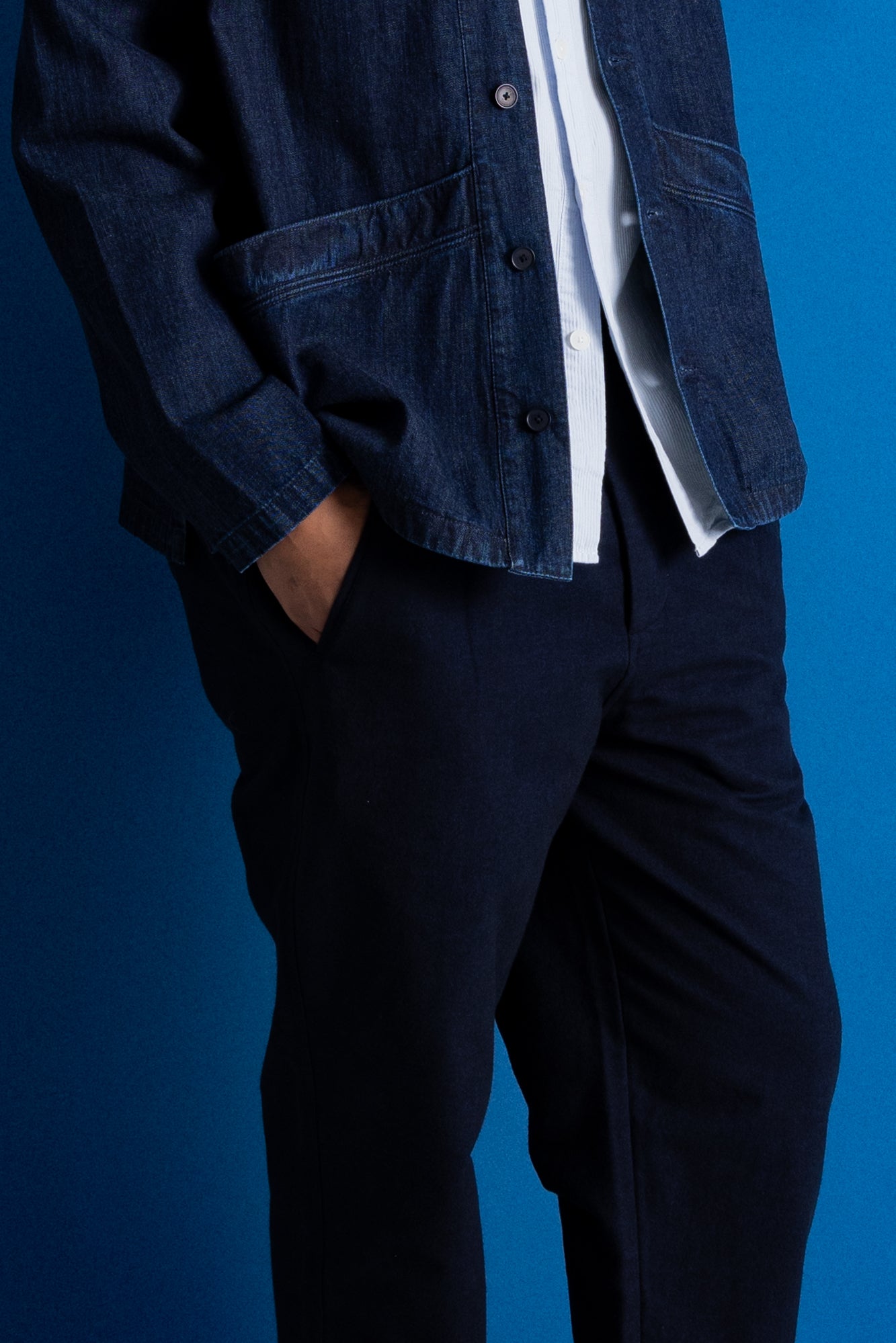 A person wearing a dark indigo denim jacket over a white shirt and dark navy trousers, standing against a solid blue background.